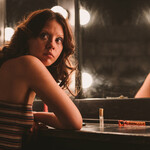 A white woman with freckles and big, curly reddish hair sits in front of a backstage vanity rimmed with bright lightbulbs. She is turned away from the mirror, towards the viewer, looking off camera with a concerned expression on her face. 