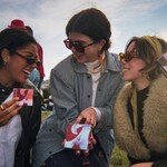 Three young women in sunglasses and coats (one Brown woman with a brown braid and two white-passing women with their brown hair pulled off their faces) hold boxes of tampons and pads in August's advertisement.