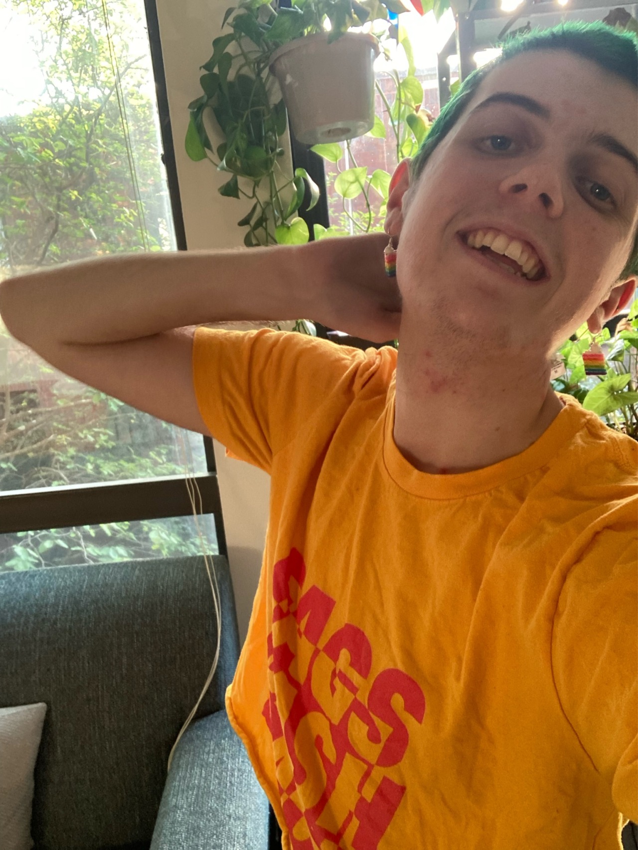 a photo of me, a white trans guy with short green hair wearing a yellow and pink crop top and standing in front of a lot of plants