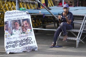 A protester drinks a cup of tea as she sits by a defaced poster carrying portraits of ousted president Gotabaya Rajapaksa, center, and his brothers at the entrance to president's office in Colombo, Sri Lanka, Friday, July 15, 2022. Protesters retreated from government buildings Thursday in Sri Lanka, restoring a tenuous calm to the economically crippled country, and the embattled president at last emailed the resignation that demonstrators have sought for months.