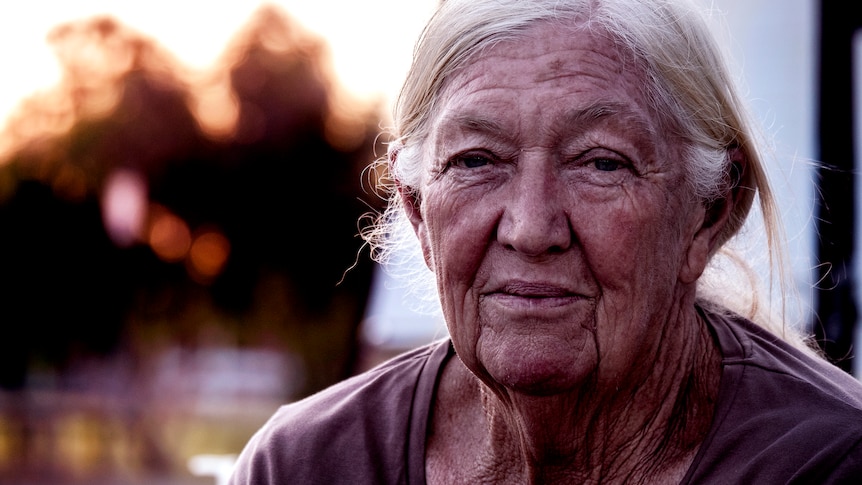 A close up photo of an elderly women with a weathered face, grey hair, blurred trees in background.