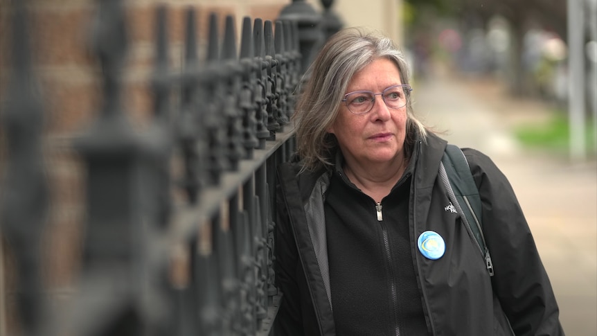 A woman with glasses wearing dark top leaning against a iron fence