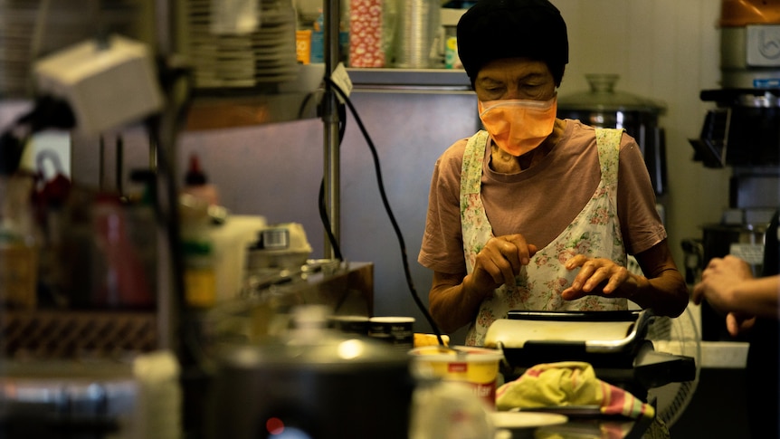 A woman in a mask and apron in a small commercial kitchen.