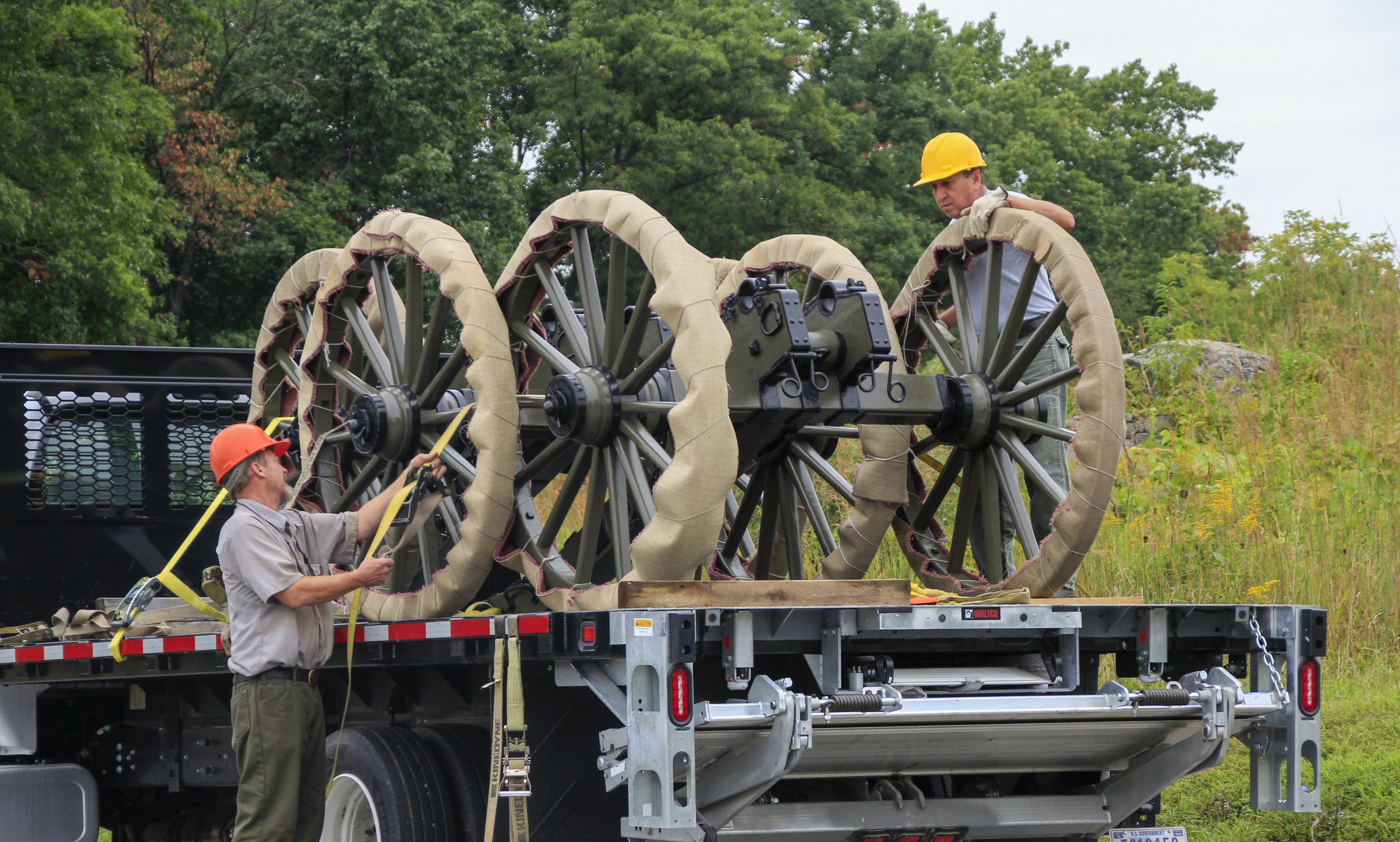 Three civil war cannon carriages are on a flat bed truck. The wheels are wrapped in brown burlap. Two national park service employees with orange and yellow hard hats prepare them to be unloaded.