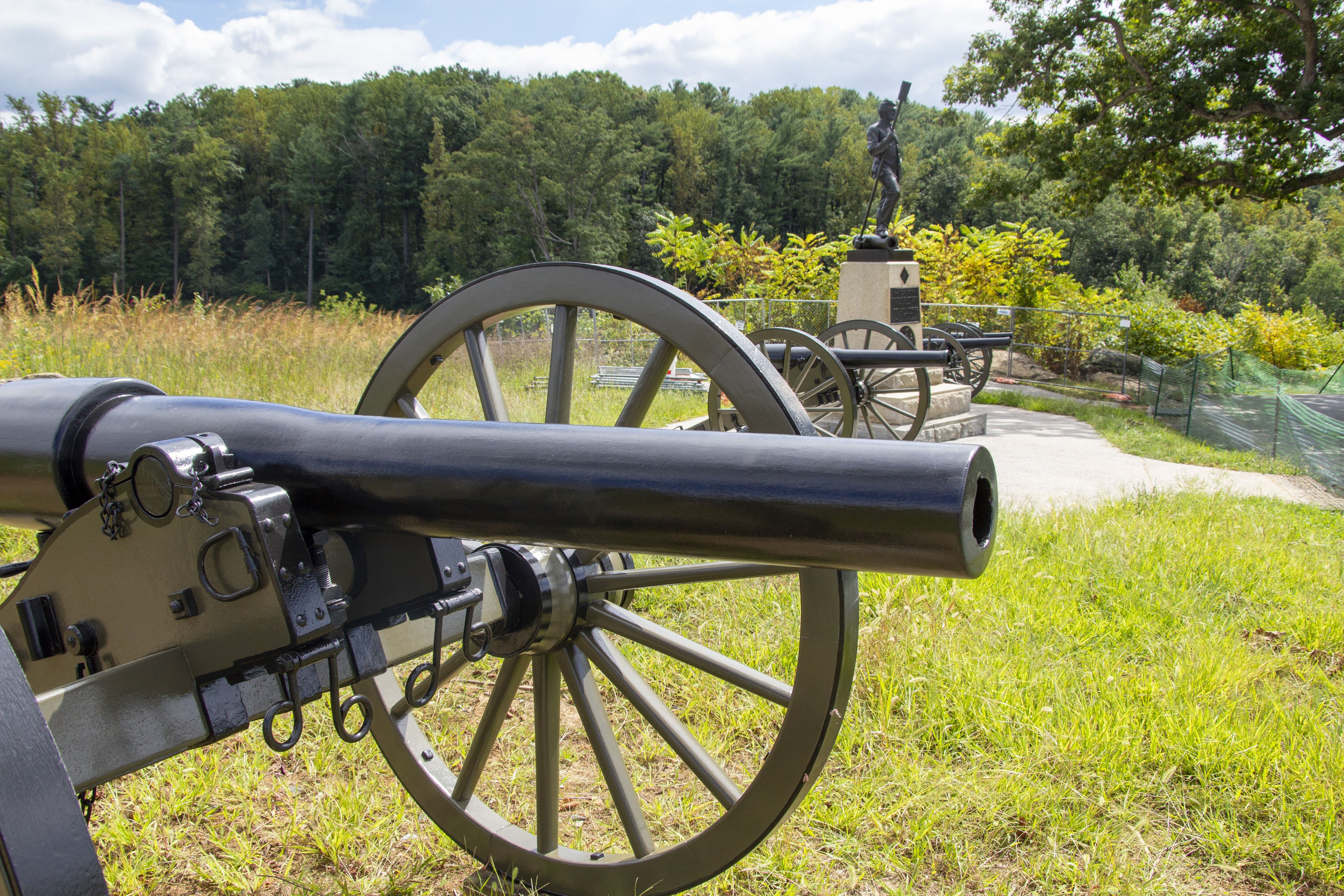 A row of four cannons sit in a line. There is a taller granite and bronze monument in the middle. The figure on the top of the monument depicts a Civil War artillery soldier. There is a heavily wooded hill in the background.