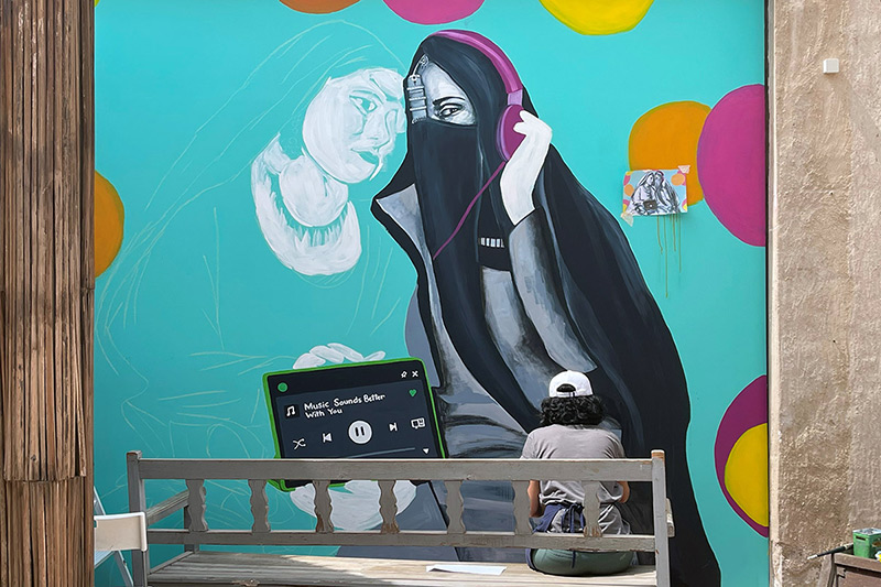 A person seated, facing away, paints a large mural depicting an Arab woman wearing head and face coverings, listening to pink headphones connected to a device playing music, on a background of bright teal with pink and orange dots.