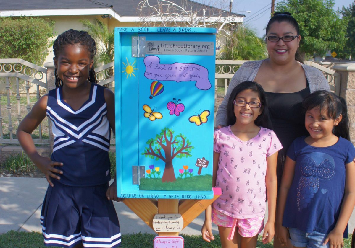 three young female children and a woman standing next to a bright blue little free library