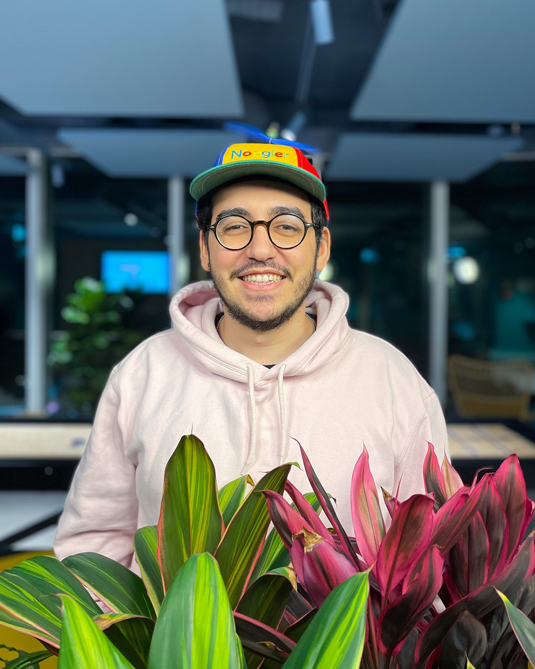 Rand wearing glasses smiling in a pink sweatshirt, noogler hat in front of plants.