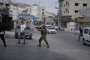 An Israeli soldier draws his weapon on Palestinians, left, as settlers, right, attack shops in Huwara, near the West Bank town of Nablus, Thursday, Oct. 13, 2022.