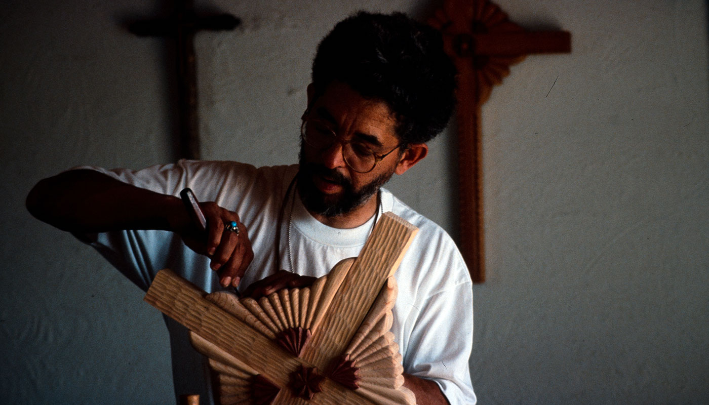 A man carves detail into a wooden cross, with other crosses hanging on the wall behind him.