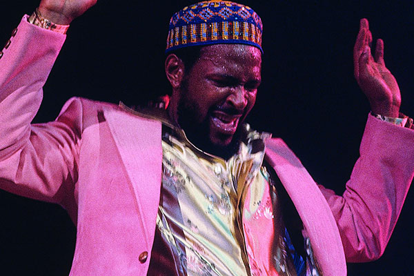 Marvin Gaye on stage in a pink jacket and silvery shirt, hands both raised, and sweat on his brow.