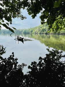 A small open boat on the calm surface of the river Chaliyar, in Kerala, India. The sky is dark with clouds, and the river banks are thick with foliage.