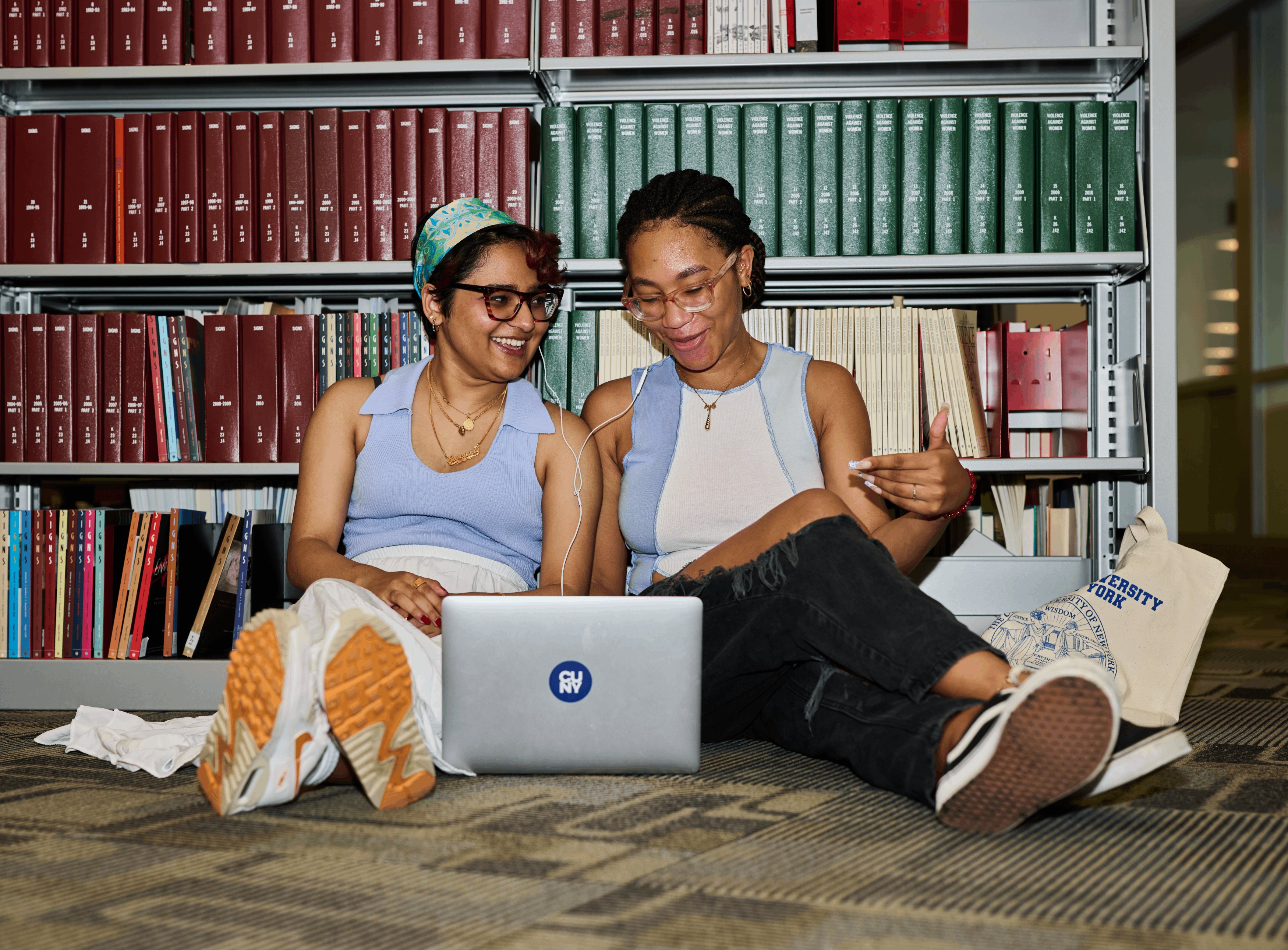 Two female students sitting on the floor of a library share headphones and laugh together.