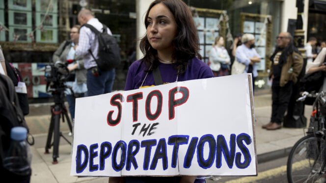 A person with long brown hair, medium-dark skin and a purple top carries a white banner on which is written in black, red and purple lettering: 'Stop the Deportations'