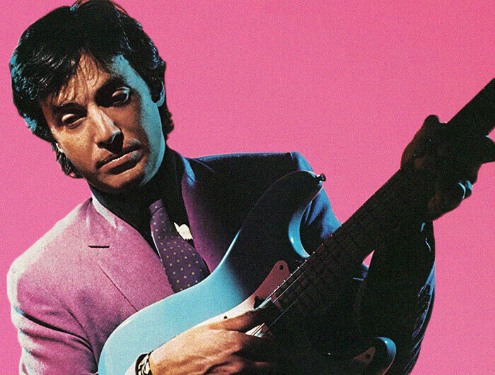 A man in a light pink suit holds a baby blue guitar.