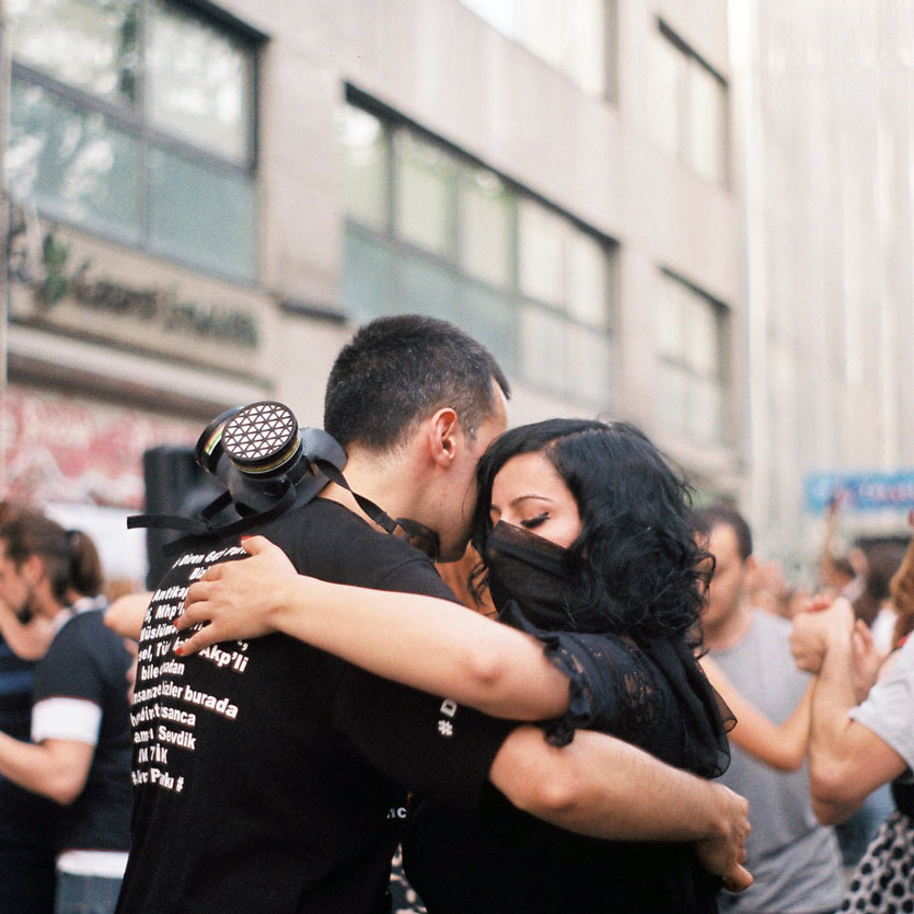 two protestors, adorned with gas masks and balaclavas, dance the tango together in the street