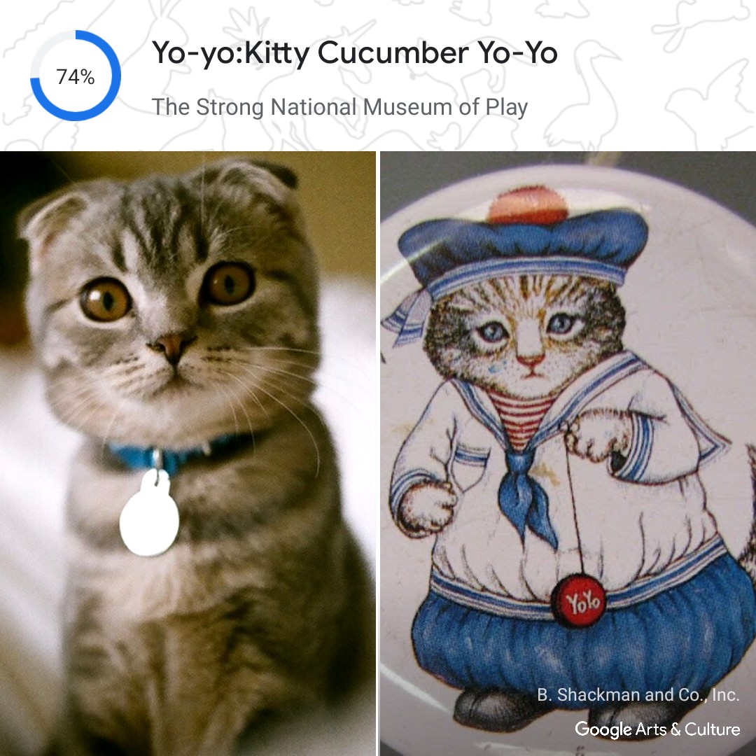 On the left, a photo of a cat with a blue collar staring at the camera. On the right, a drawing of a cat wearing a blue and white sailor outfit. Above the cats, the title of the artwork is displayed: Yo-yo:Kitty Cucumber Yo-Yo, from The Strong National Museum of Play. It is from the Google Arts & Culture app, and they have a 74% match. At the bottom right, it says: B. Shackman and Co., Inc. Google Arts & Culture