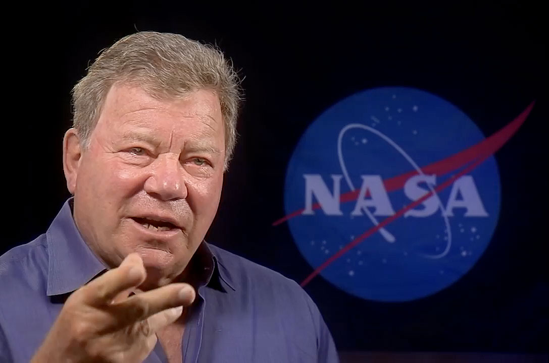 Stillshot of William Shatner talking and gesticulating with his hands in front of a dark screen that shows the NASA meatball dimly lit.