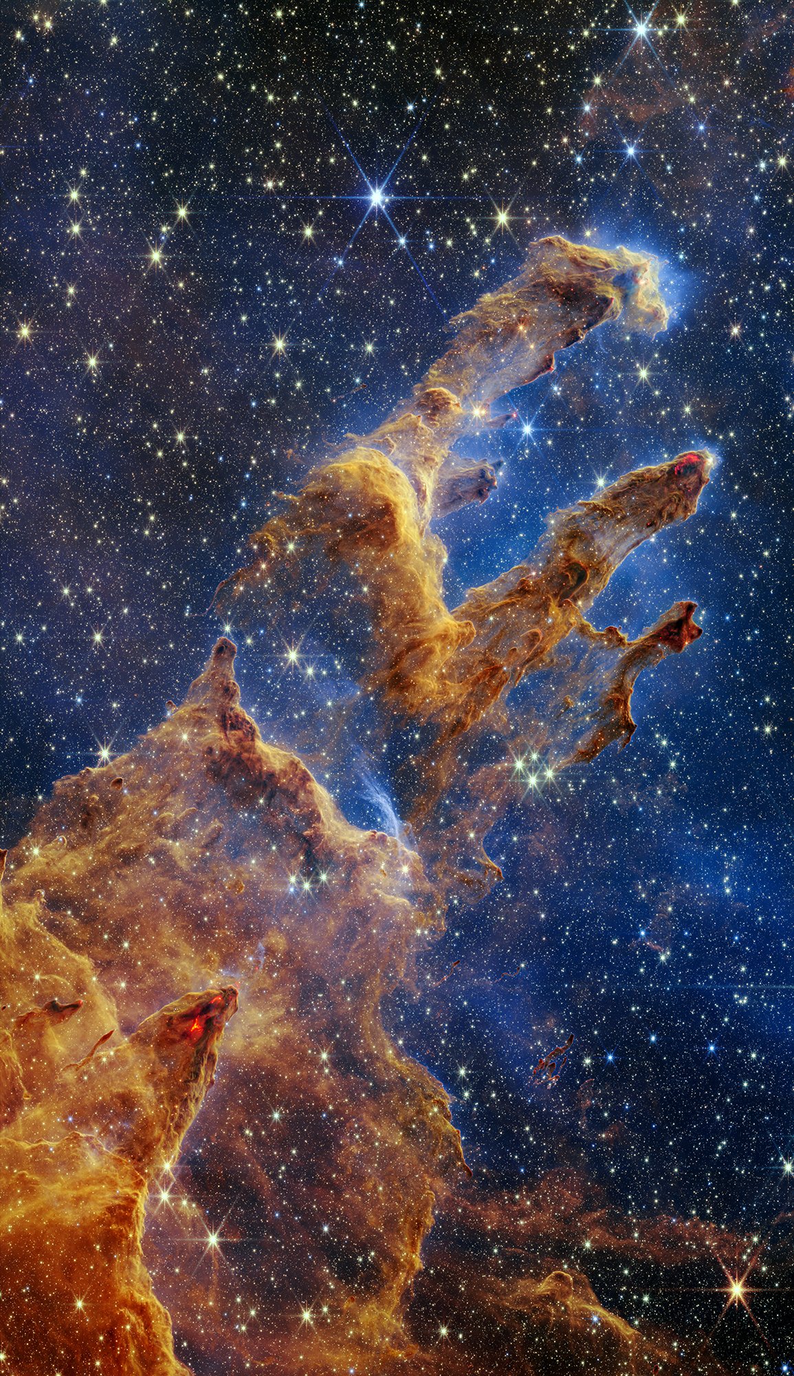 This Webb image of the “Pillars of Creation” has layers of semi-opaque, rusty red gas and dust that start at the bottom left and go toward the top right. There are three prominent pillars rising toward the top right. The left pillar is the largest and widest. The peaks of the second and third pillars are set off in darker shades of brown and have red outlines. Peeking through the layers of gas and dust is the background, set in shades of blue and littered with tiny yellow and blue stars. Many of the tips of the pillars appear tinged with what looks like lava. There are also tiny red dots at the edges of the pillars, which are newly born stars. Credits: NASA, ESA, CSA, STScI; Joseph DePasquale (STScI), Anton M. Koekemoer (STScI), Alyssa Pagan (STScI)