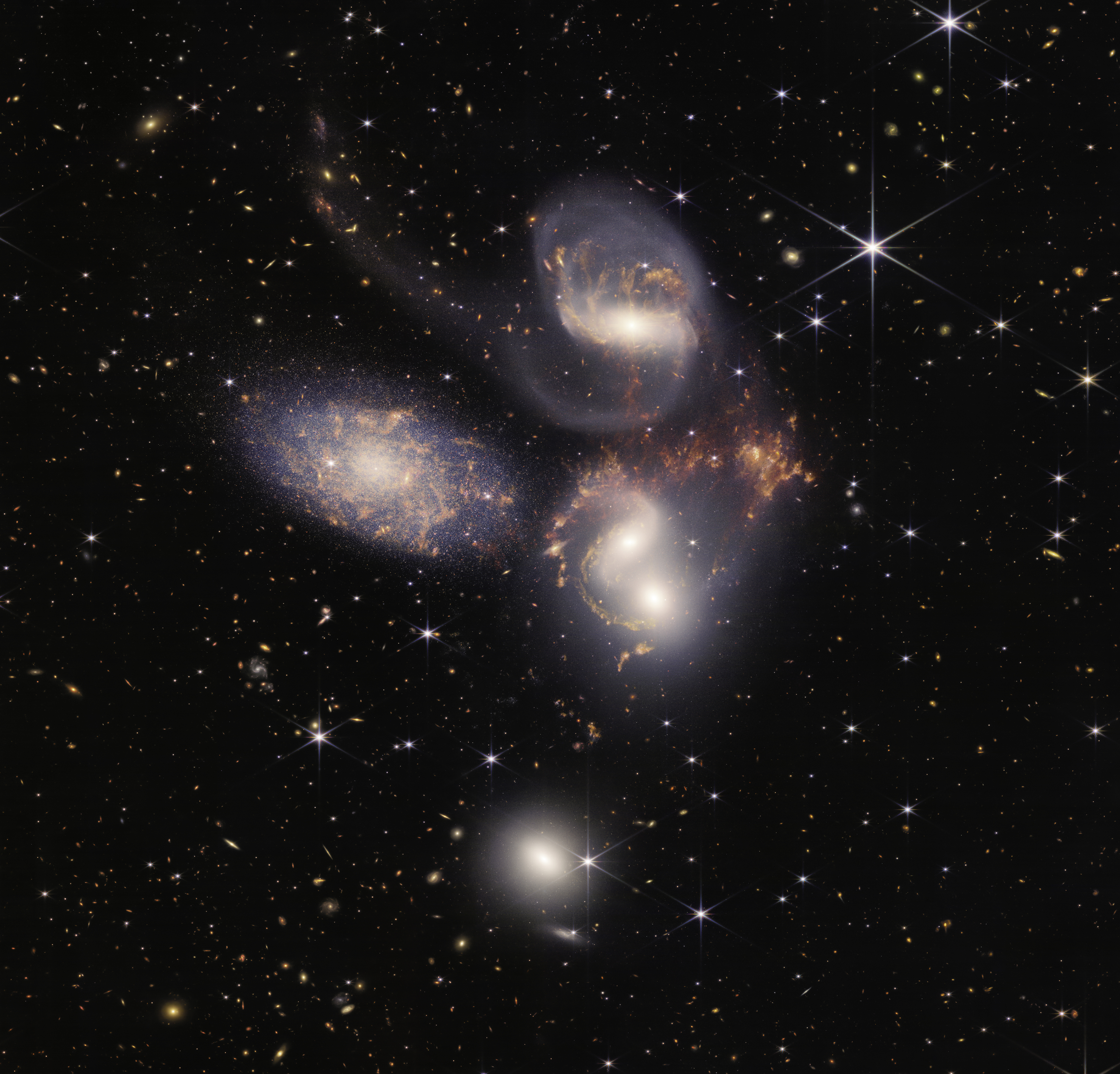 A group of five galaxies that appear close to each other in the sky: two in the middle, one toward the top, one to the upper left, and one toward the bottom. Four of the five appear to be touching. One is somewhat separated. In the image, the galaxies are large relative to the hundreds of much smaller (more distant) galaxies in the background. All five galaxies have bright white cores. Each has a slightly different size, shape, structure, and coloring. Scattered across the image, in front of the galaxies are number of foreground stars with diffraction spikes: bright white points, each with eight bright lines radiating out from the center.