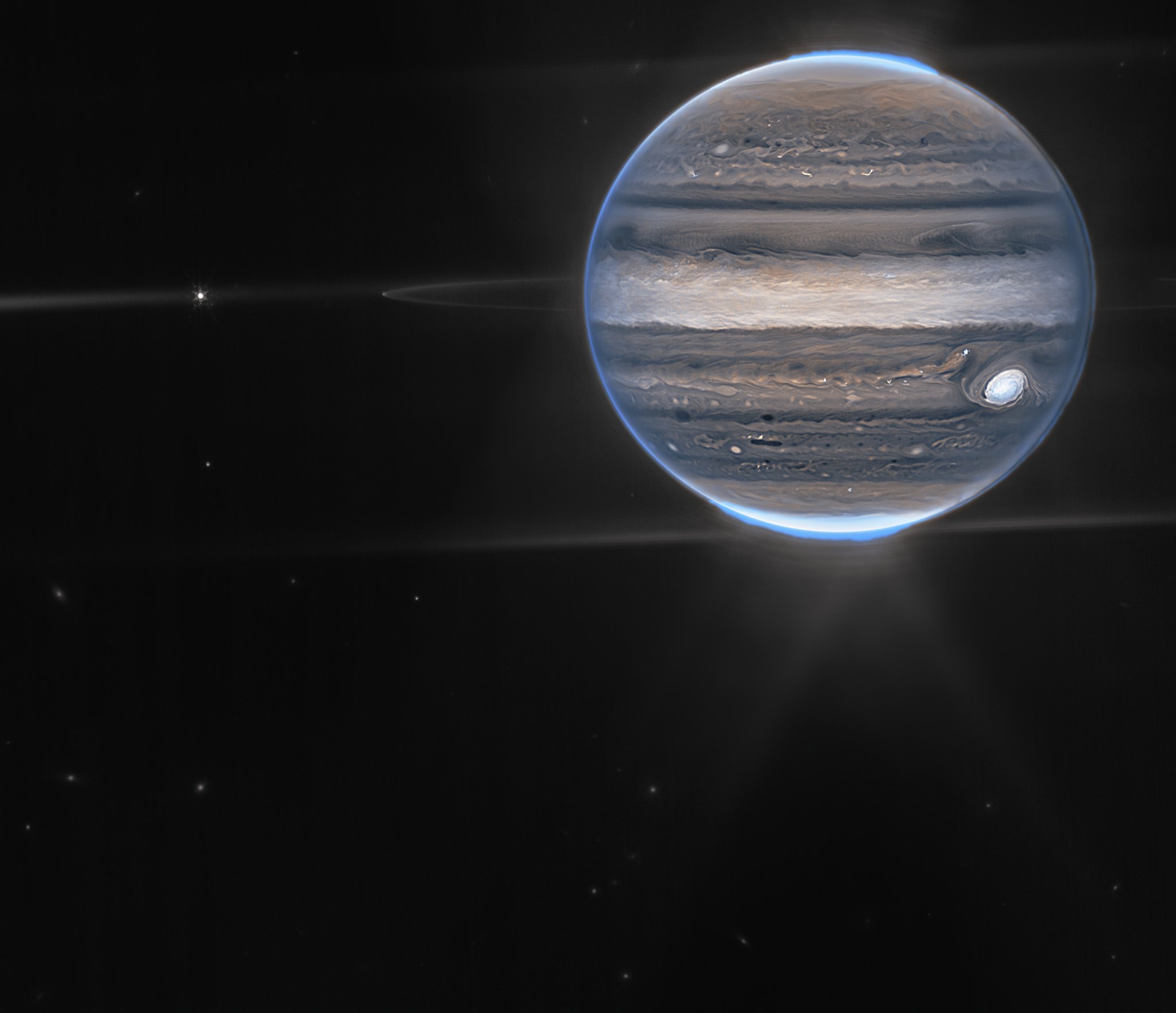 A wide field view showcases Jupiter in the upper right quadrant. The planet’s swirling horizontal stripes are rendered in blues, browns, and cream. Electric blue auroras glow above Jupiter’s north and south poles. A white glow emanates out from the auroras. Along the planet’s equator, rings glow in a faint white. These rings are one million times fainter than the planet itself! At the far left edge of the rings, a moon appears as a tiny white dot. This moon is only about 12 miles (20 km) across. Slightly further to the left, another moon, about 100 miles (150 km) across, glows with tiny white diffraction spikes. The rest of the image is the blackness of space, with faintly glowing white galaxies in the distance.