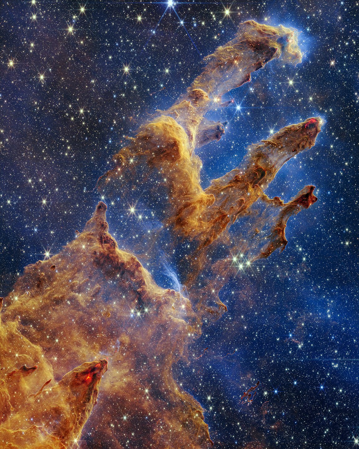 This Webb image of the “Pillars of Creation” has layers of semi-opaque, rusty red gas and dust that start at the bottom left and go toward the top right. There are three prominent pillars rising toward the top right. The left pillar is the largest and widest. The peaks of the second and third pillars are set off in darker shades of brown and have red outlines. Peeking through the layers of gas and dust is the background, set in shades of blue and littered with tiny yellow and blue stars. Many of the tips of the pillars appear tinged with what looks like lava. There are also tiny red dots at the edges of the pillars, which are newly born stars.