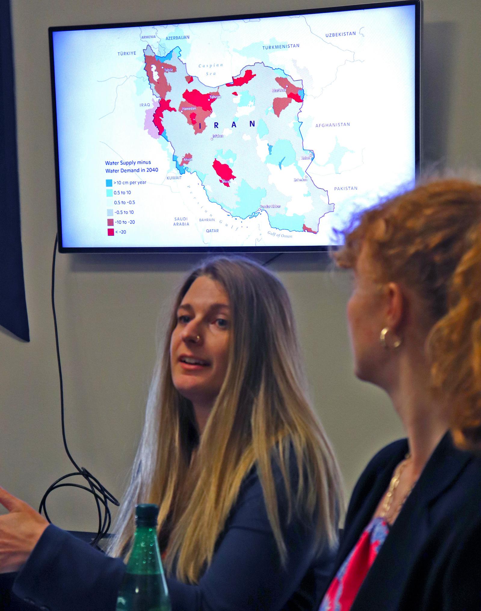 A woman speaks in front of a screen displaying a map of Iran with colored polygons