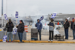 Workers six workers seen from far away hold picket signs by a burn barrel.