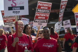 a group of smiling nurses, mostly African American, in red shirts hold signs saying ‘patients before profits’