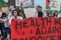 A group of people in white medical coats stand behind a red banner reading ‘Health Workers Against Genocide in Gaza.’