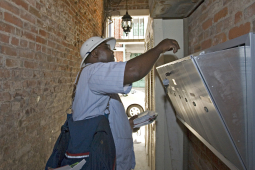 A letter carrier with a big shoulder bag puts mail in a grouped mailbox in an alley, he is backlit.