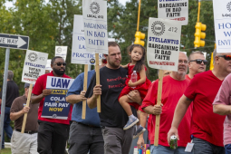 A group of people in red shirts, one man carrying a child march past the camera with signs saying Stellantis is barganing in bad faith.