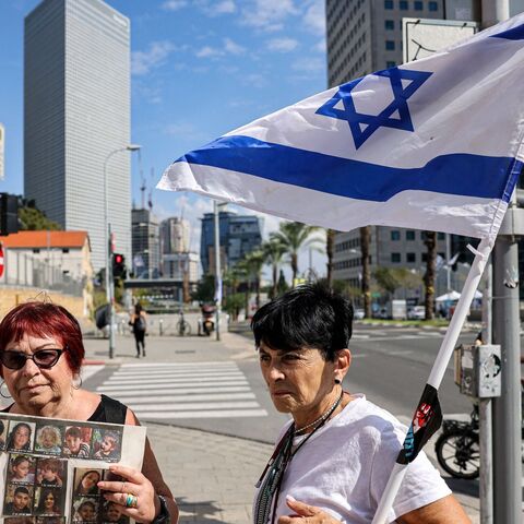 An elderly woman stands with an Israeli flag next to another holding a sign showing the faces of young Israeli hostages held by Palestinian militants since the October 7 attack near Azrieli Mall in Tel Aviv on October 18, 2023. (Photo by AHMAD GHARABLI / AFP) (Photo by AHMAD GHARABLI/AFP via Getty Images)