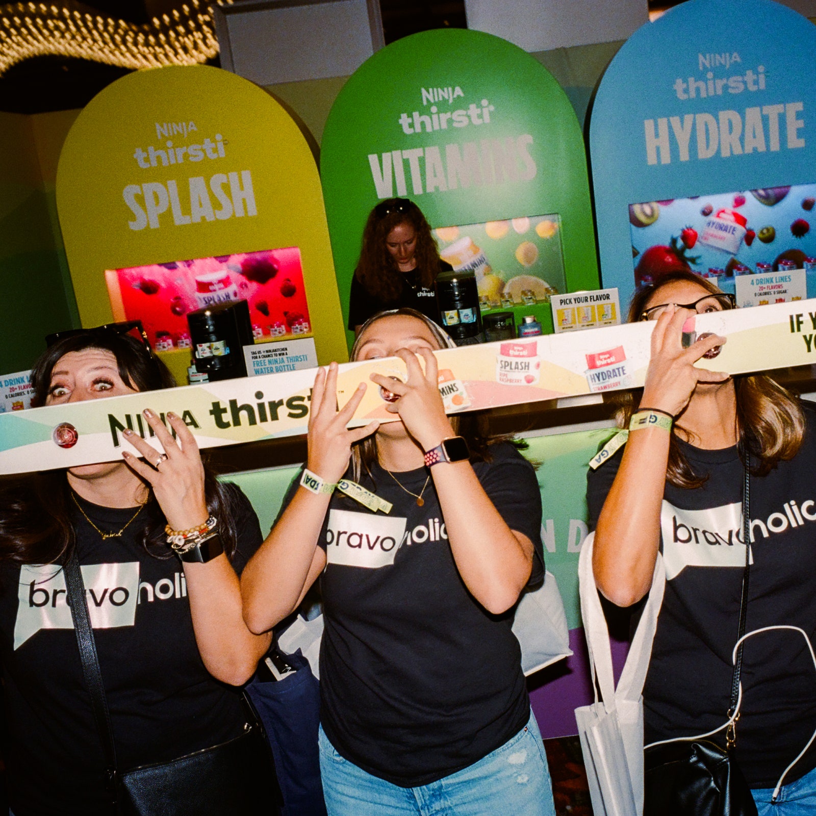 Three people stand side by side and raise a shotski to their mouths. All are wearing black tshirts that say bravoholic...