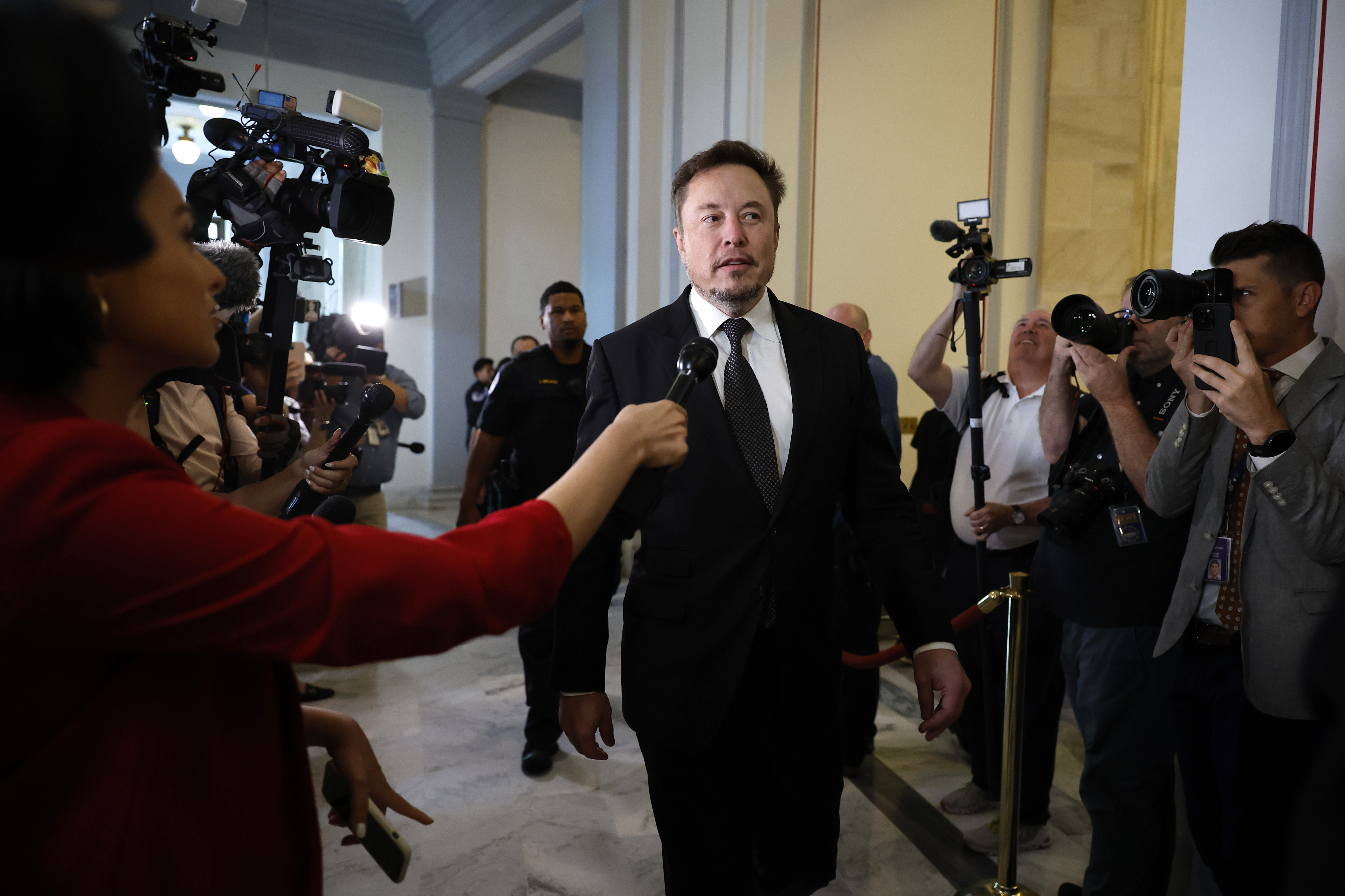 Elon Musk, wearing a black suit and white shirt, speaks to a row of reporters with microphones and cameras as he walks down a hallway.