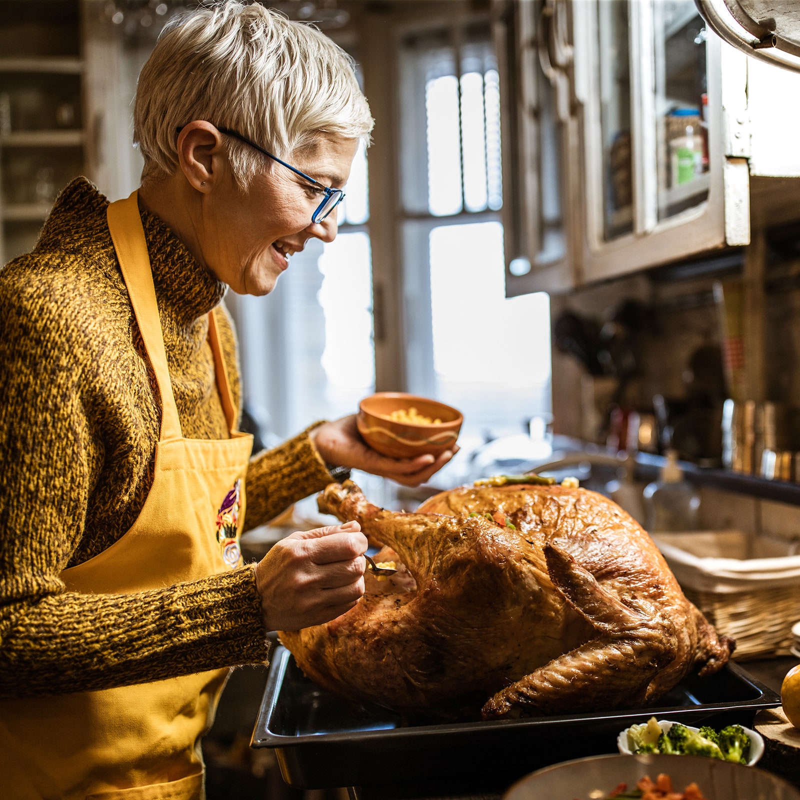 A person cooking a Thanksgiving turkey in a kitchen. The person has short white hair wears glasses is wearing a yellow...