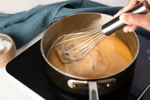 Roux whisked in a saucepan on an induction stove, and in the surroundings, a bowl of flour and a blue kitchen towel