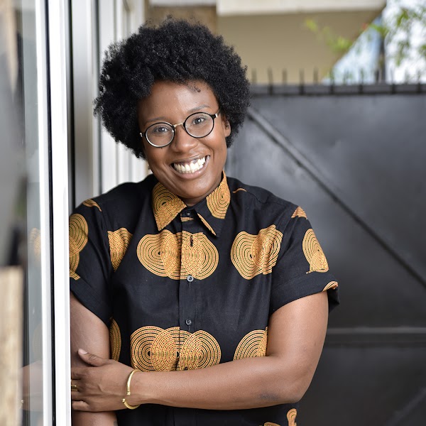 A woman with dark afro hair and circular glasses smiles while leaning against a large window pane.