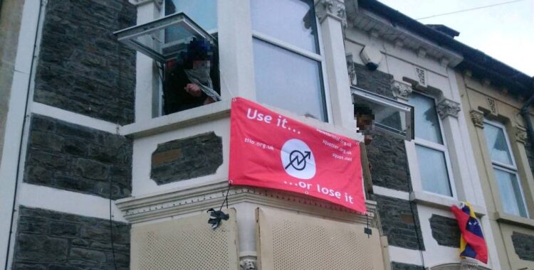 Two masked (and pixilated) people lean out of upstairs windows. Between them is a red flag with the squatting symbol that reads: Use it or loose it, squat.net