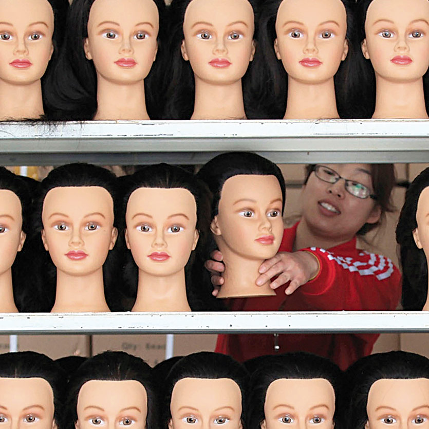 a woman of asian ethnicity works as a mannequin factory stocking a shelves that are full of caucasian mannequin heads