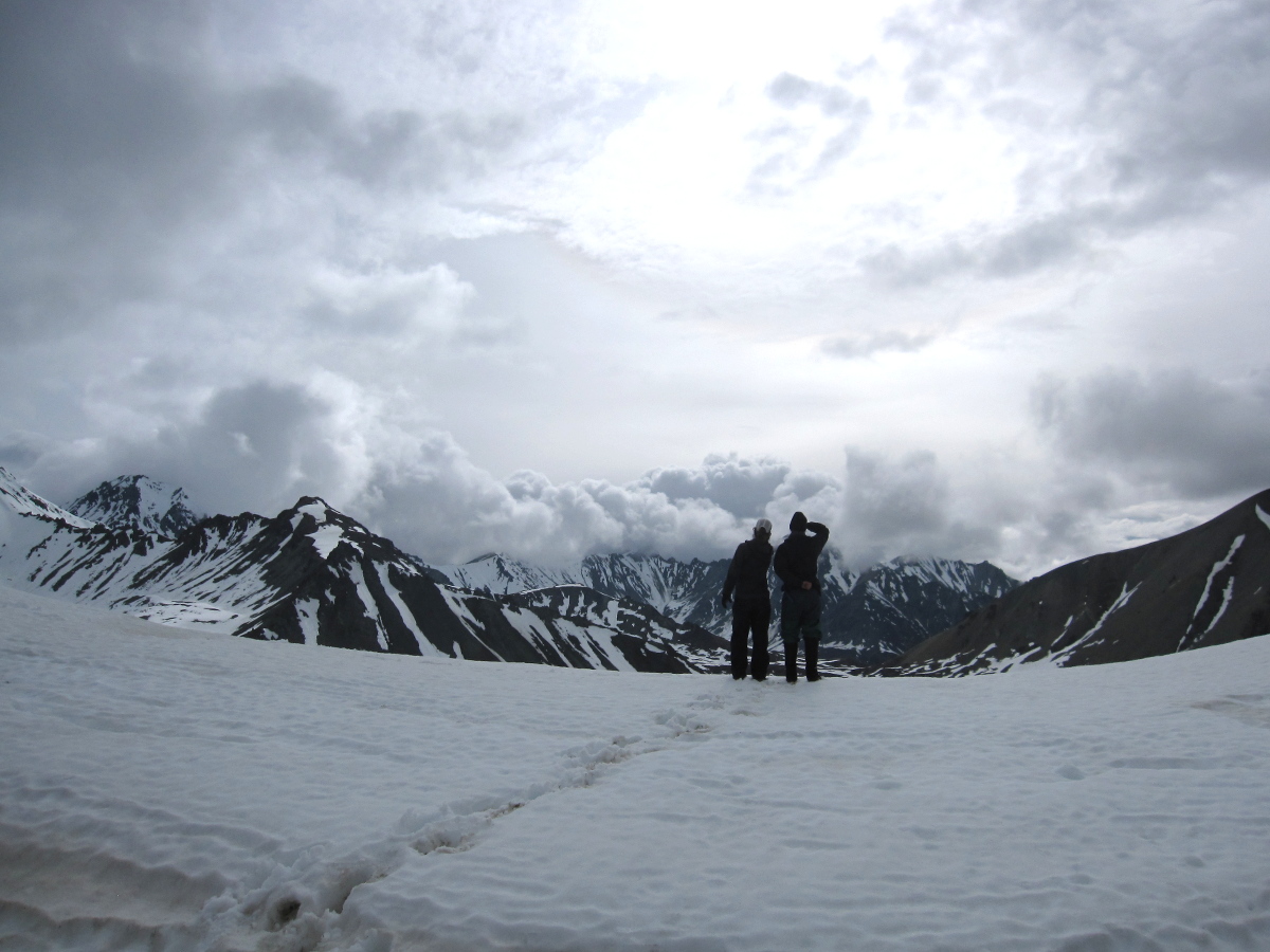 two silhouetted figures stand on a snowfield in Alaska, looking out toward mountains topped with clouds