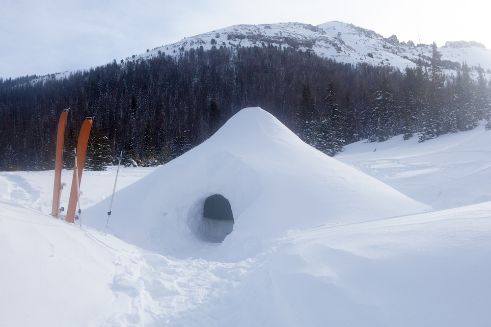 A quinzhee stands in deep snow in front of a mountain while a pair of skis rests outside its entrance.