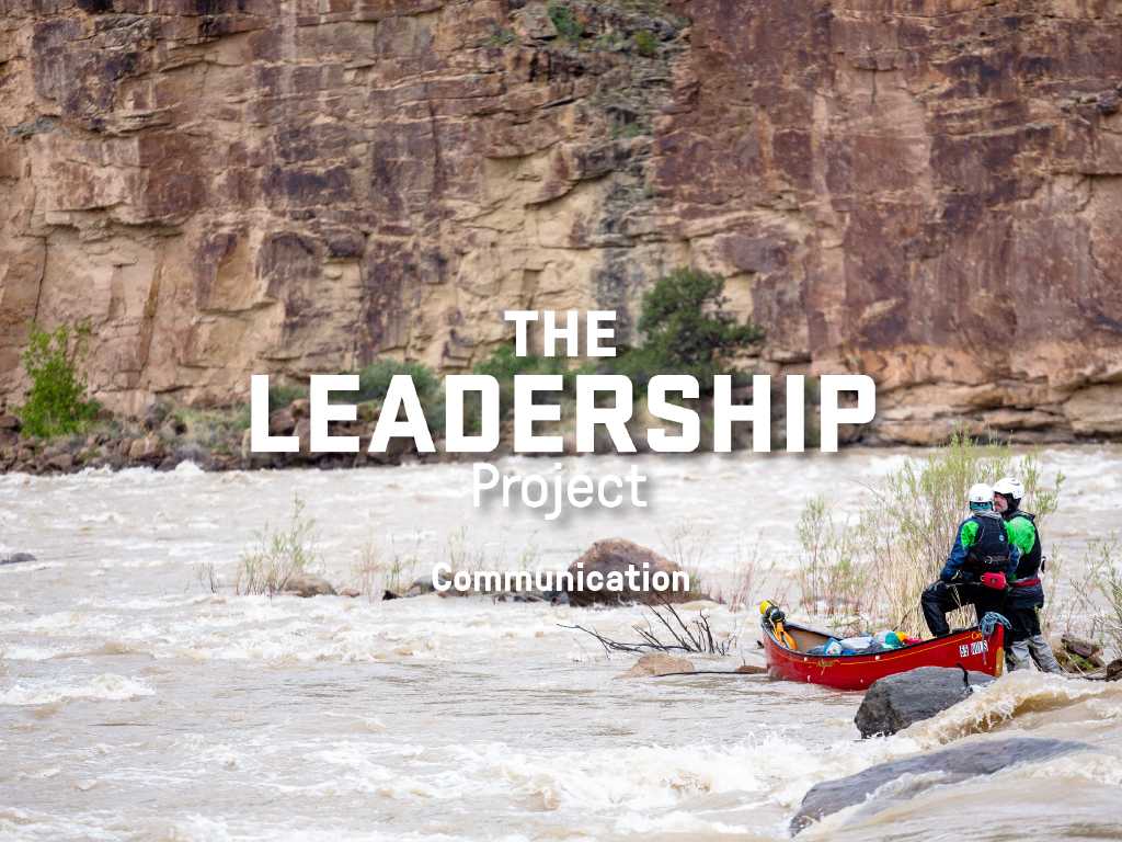 Two people talk next to a canoe on the short of a fast-moving river