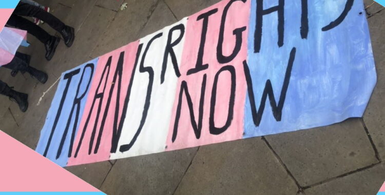 a banner painted blue pink and white in trans flag style, with black text reading TRANS RIGHTS NOW, it had been laid on the ground in front of some protesters, a raindbow gay pride flag can be seen flying behind it