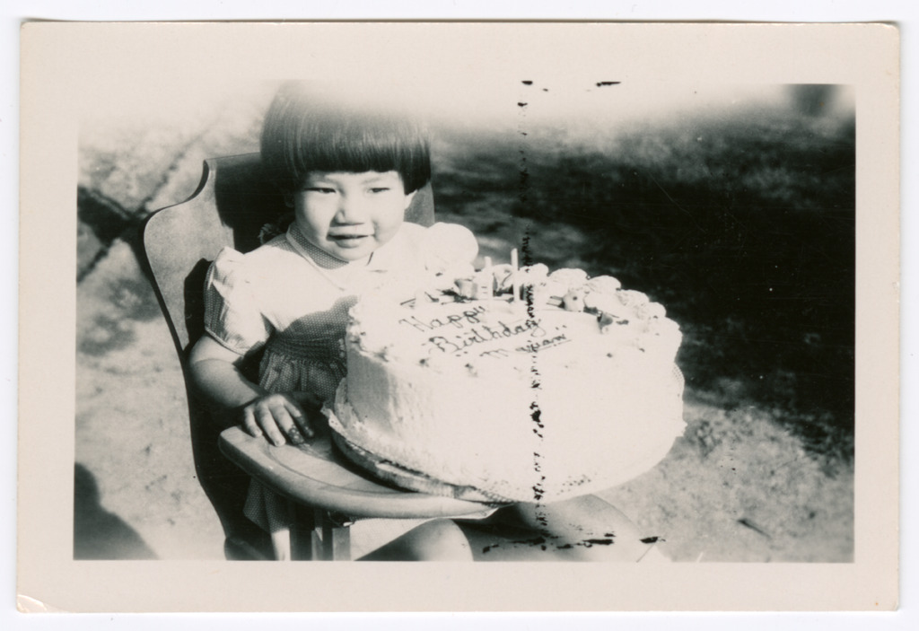 Black and white photograph of a toddler in a high chair with a large birthday cake with two candles that reads 