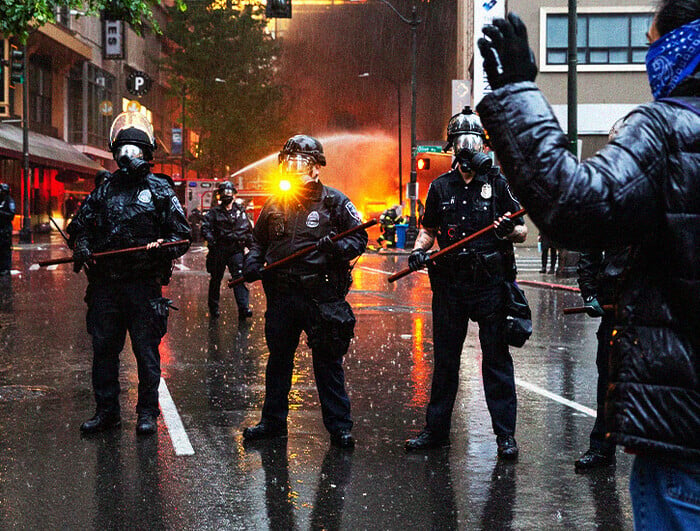 Three police officers stand in a street before a protester with their arms thrown up. In the distance, a fire.