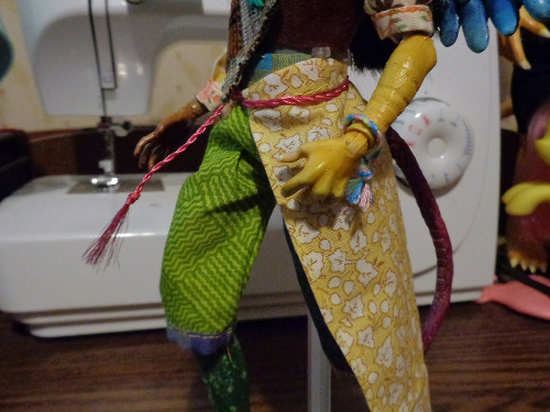 A close up of a custom doll outfit. Two toned pants in green and grey, as well as a long piece that suggests a half skirt, tied around the doll's waist. The doll's arm is visible, with a tiny friendship bracelet on the wrist.