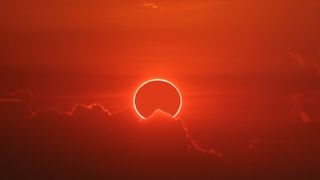 Solar eclipse photographed in Chon Buri, Thailand. The red hue image shows a distinct 'ring of fire' glowing in the sky, slightly obscured by a cloud.