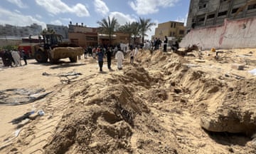 Sandy soil with large empty trench and digger in background and people in white PPE.