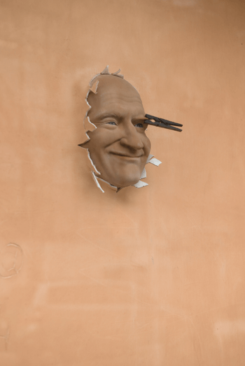 A photo of a beige sculpture where Robin Williams’s head sticks out of a wall, with a knife in his eye.